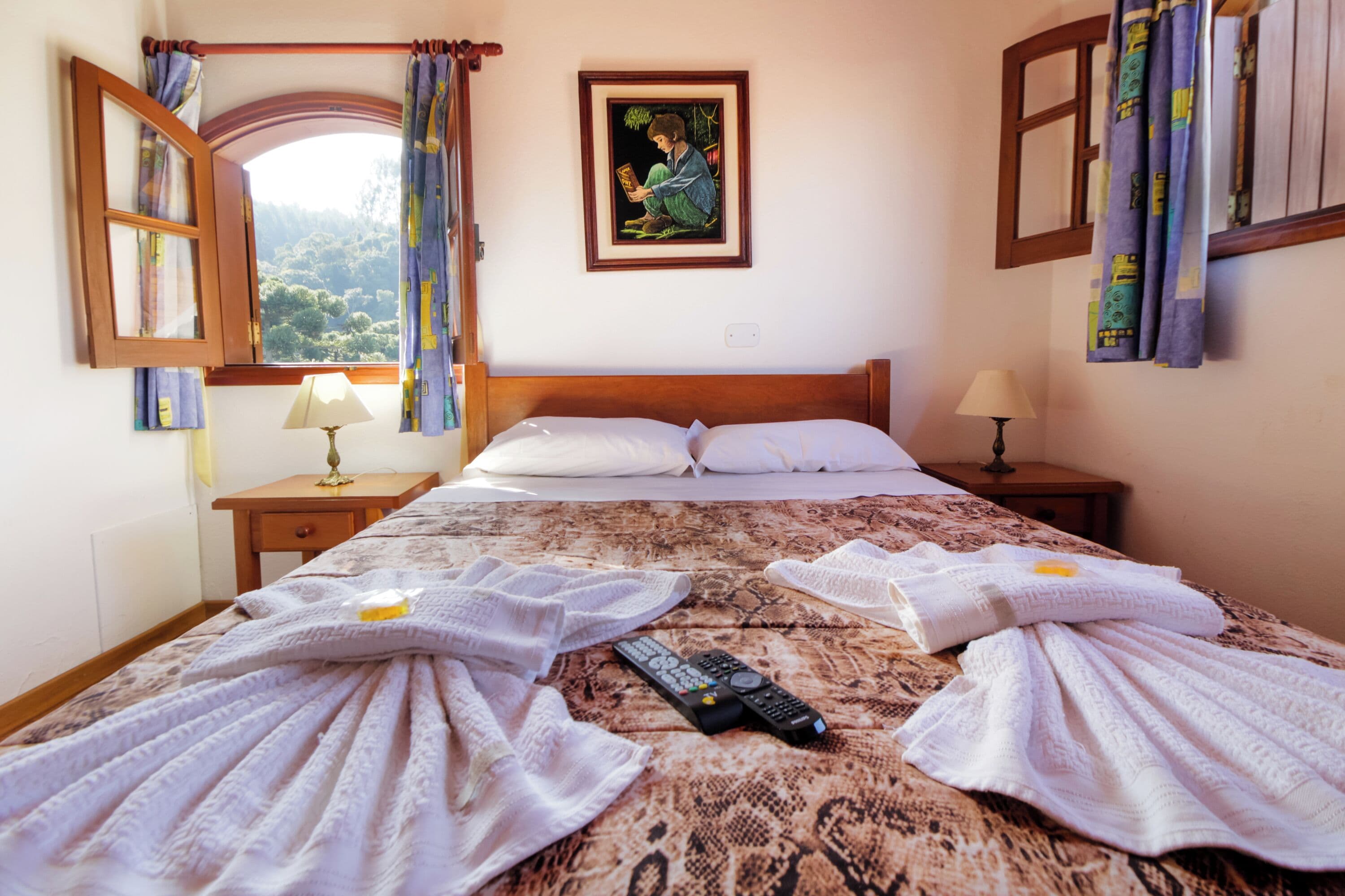 A hotel room with a bed, two bedside tables, a window with curtains, a painting on the wall, and a remote control on the bed.
