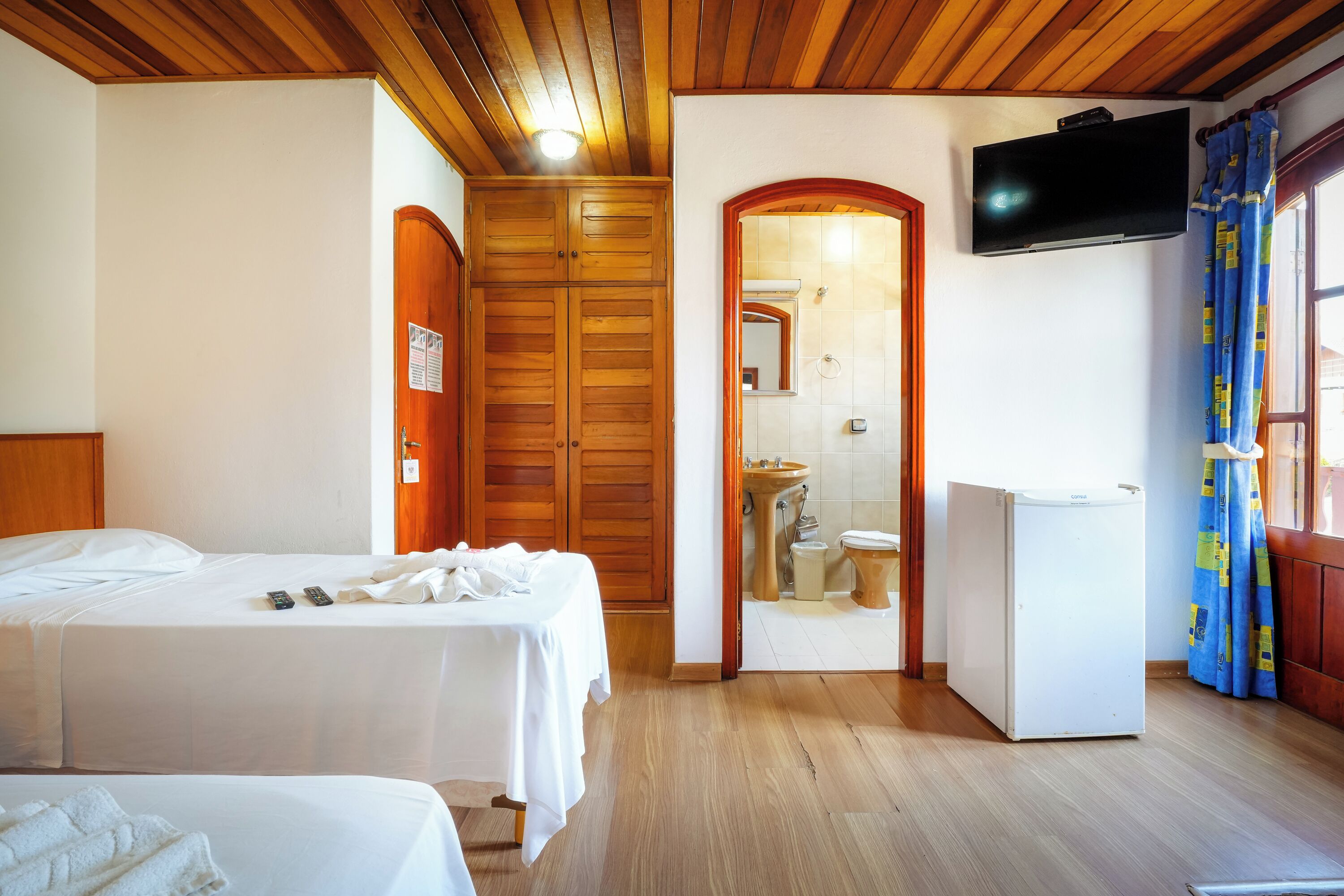 A hotel room with two beds, a wooden ceiling, a TV, a small refrigerator, and a bathroom visible through an arched doorway.