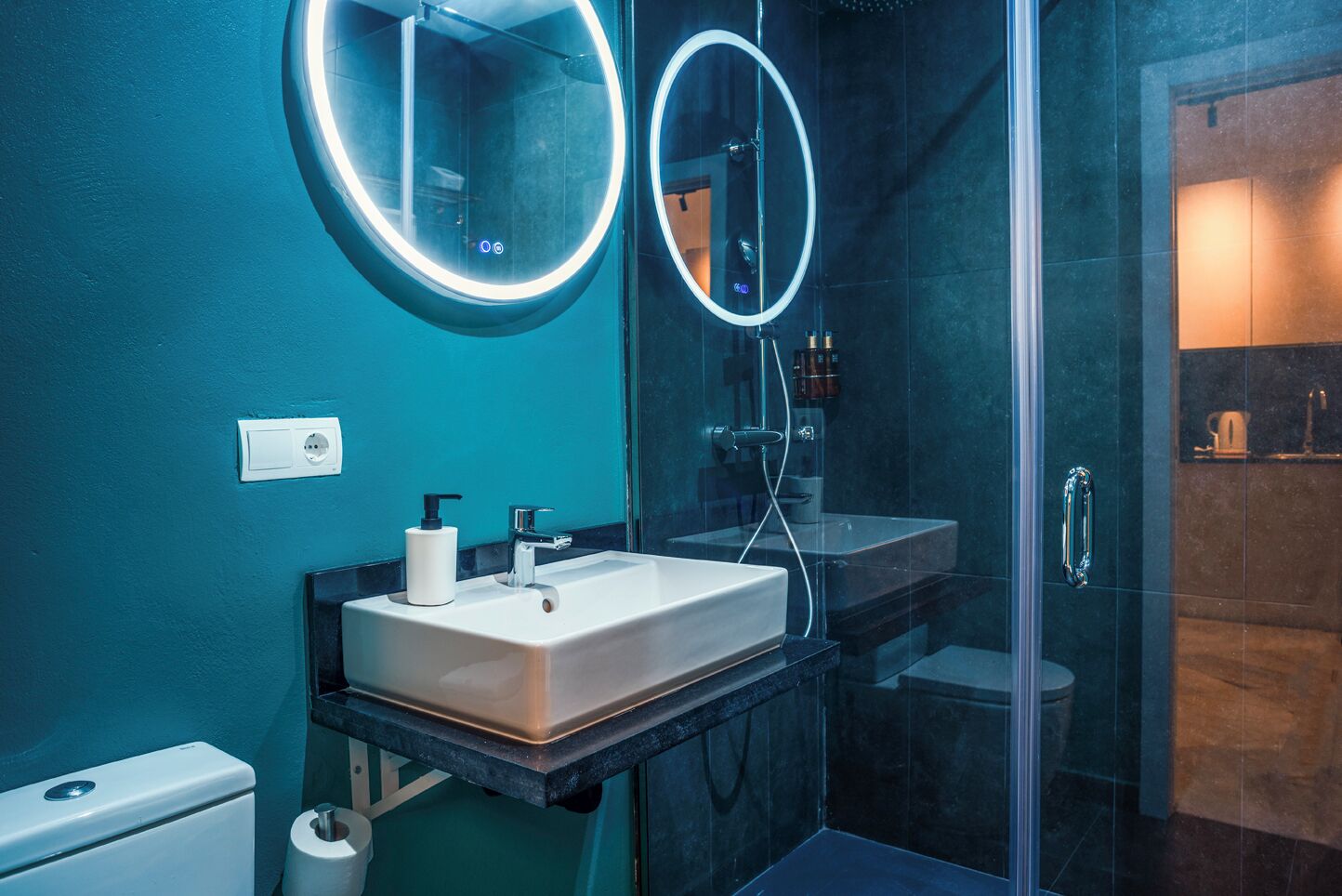 A modern bathroom with a glass shower enclosure, a white sink with a soap dispenser, and a mirror with a circular light.