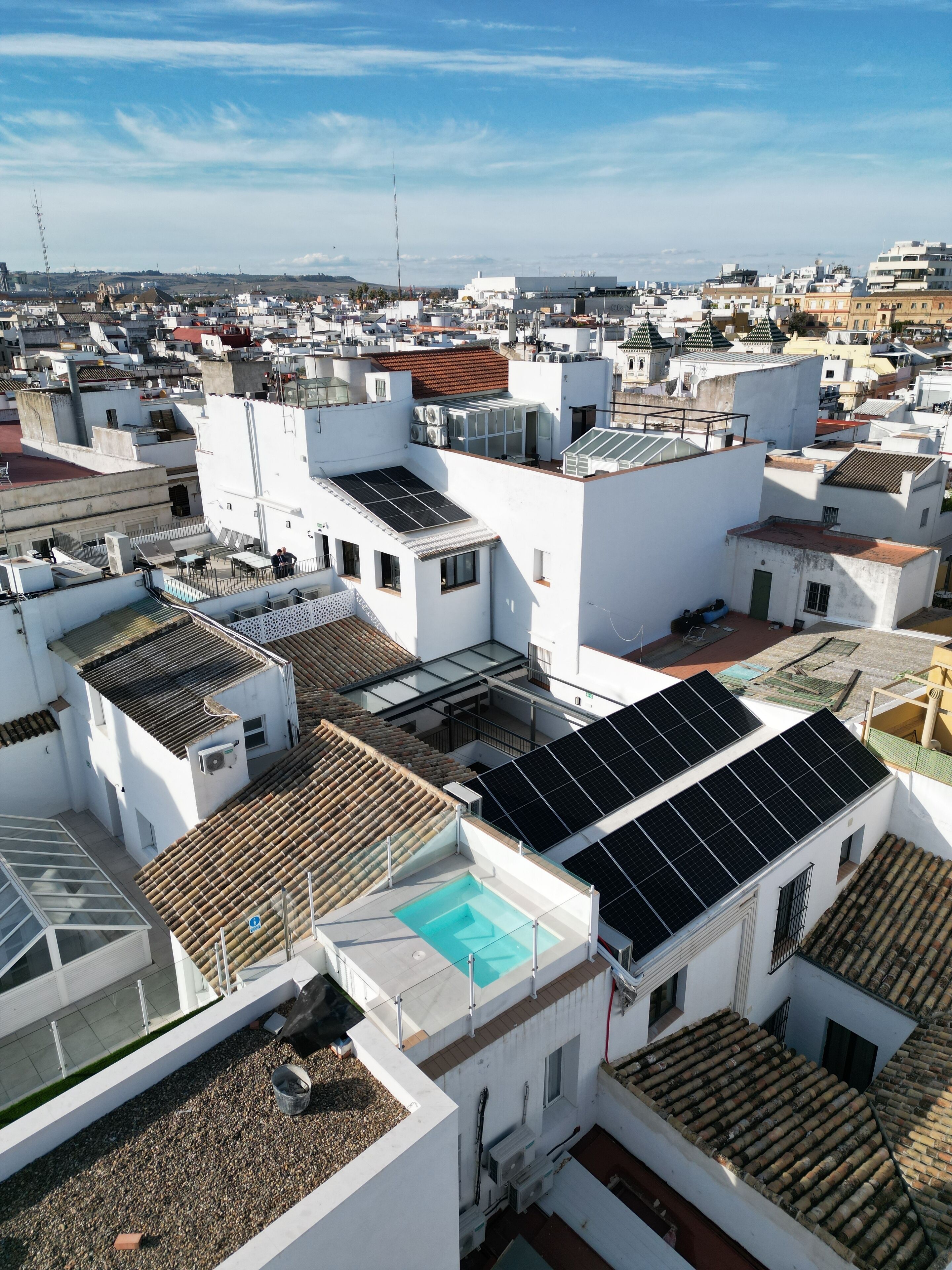 A rooftop with solar panels, a swimming pool, and multiple buildings with white walls and red-tiled roofs.