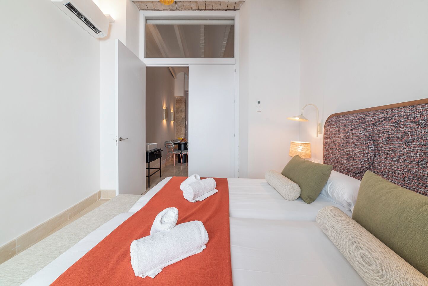 A hotel room with a bed, a sofa, a desk, and a dining area visible through an open door.