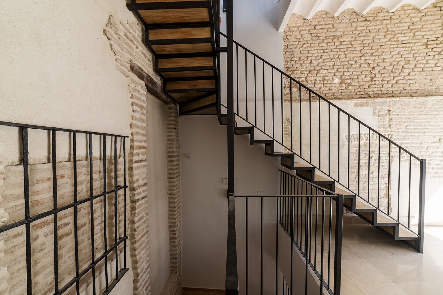 Un escalier avec des rampes noires et des marches en bois, menant à une pièce dont un mur est en briques et le plafond est blanc.