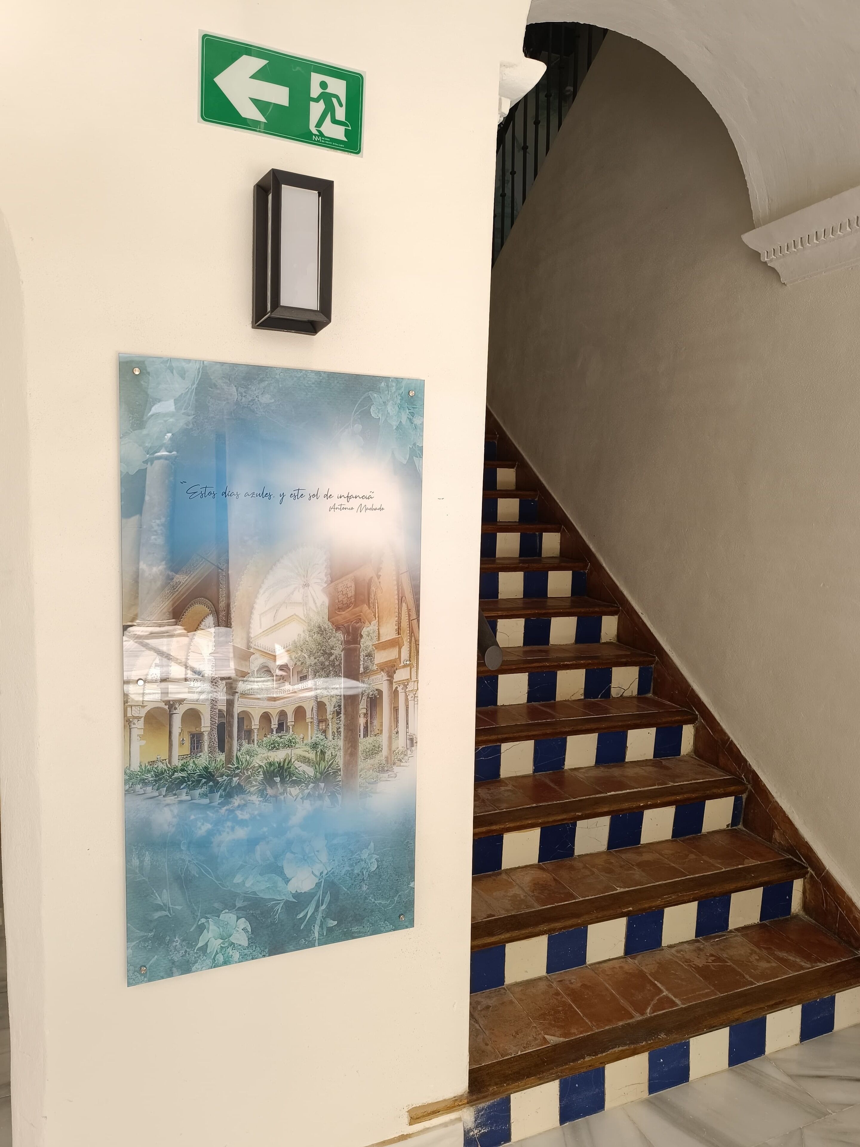 A staircase with blue and white striped steps, an emergency exit sign, and a framed picture of a building with text.