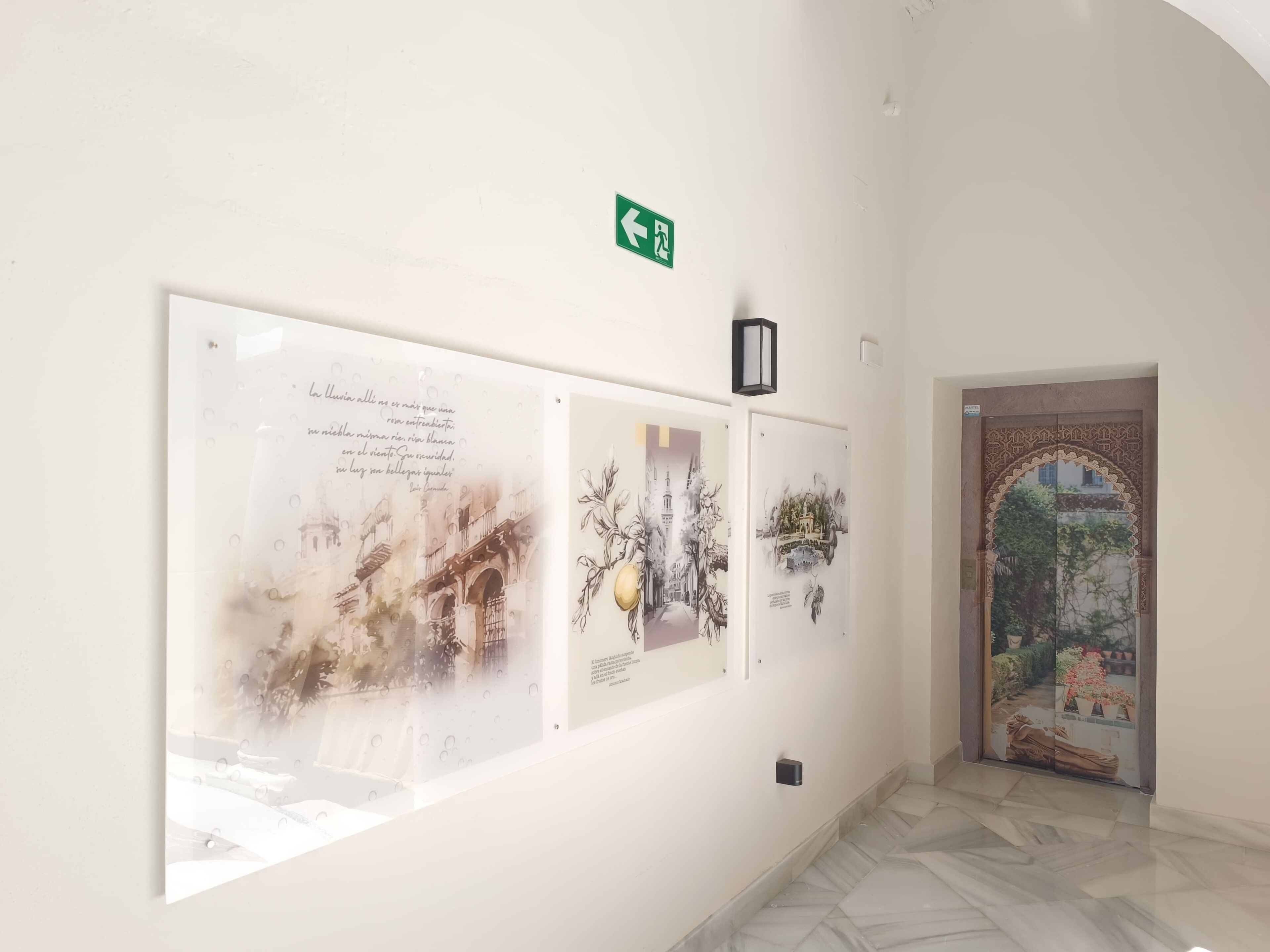 A hallway with three framed artworks on the wall, a green exit sign, and a door with intricate design.