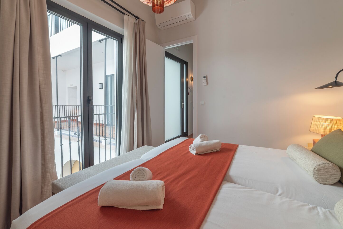 Une chambre d’hôtel avec un grand lit, deux serviettes enroulées, une couverture rouge, une table de chevet avec une lampe et une vue sur le balcon extérieur.