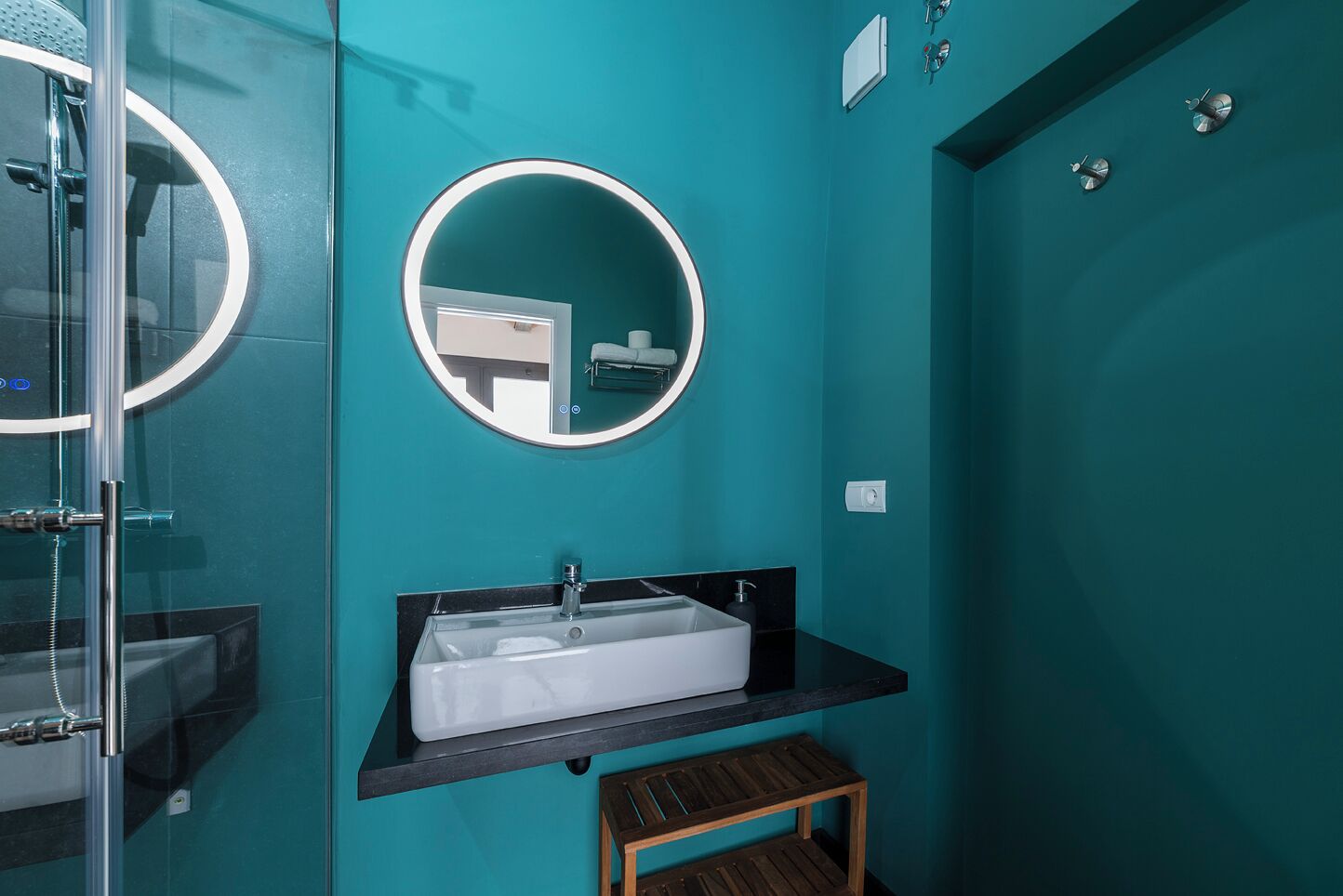 Un baño con pared color turquesa, espejo redondo, lavabo y ducha.