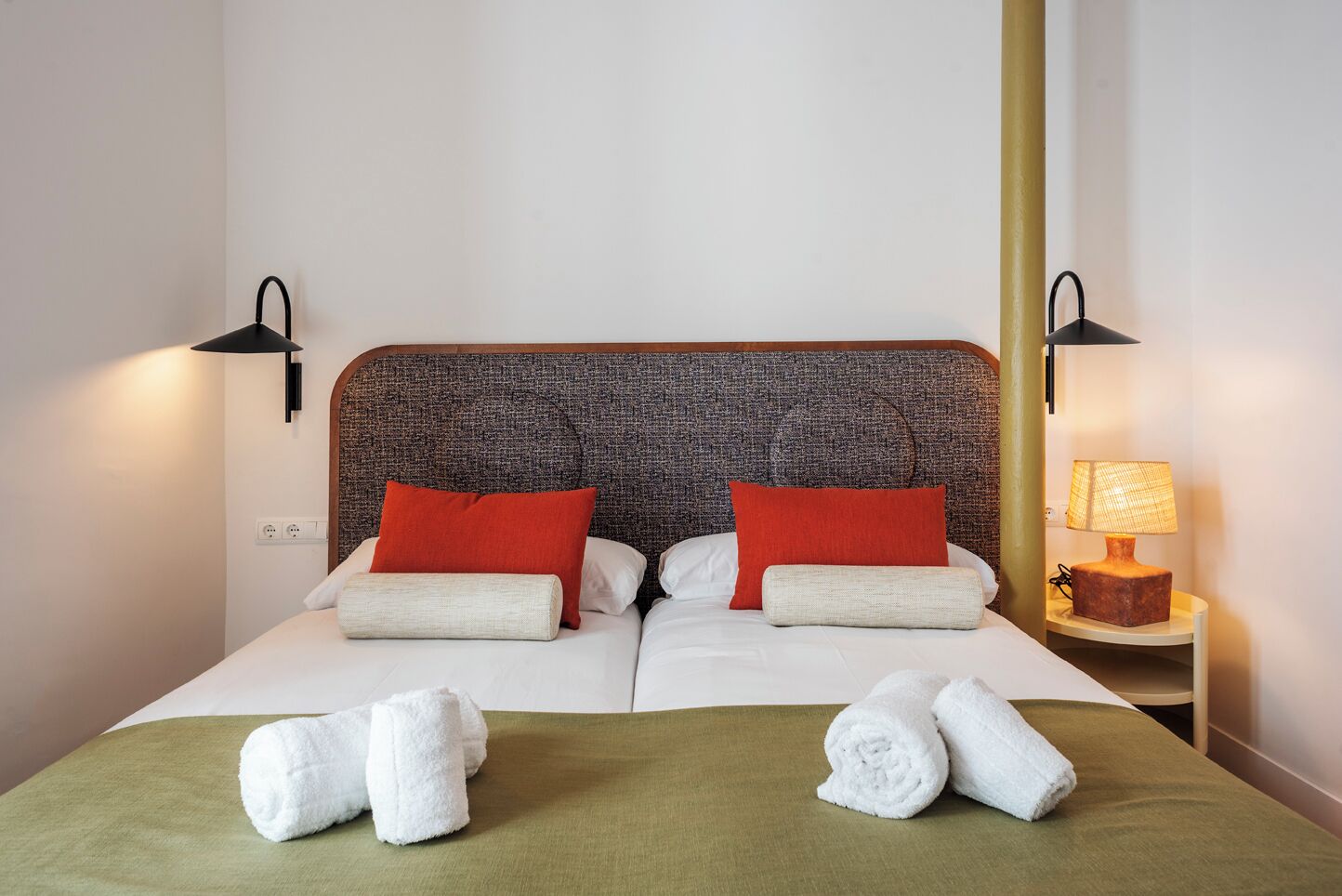 A double bed with two red pillows, a green bedspread, and two rolled white towels. There are two wall-mounted lamps on either side of the bed.