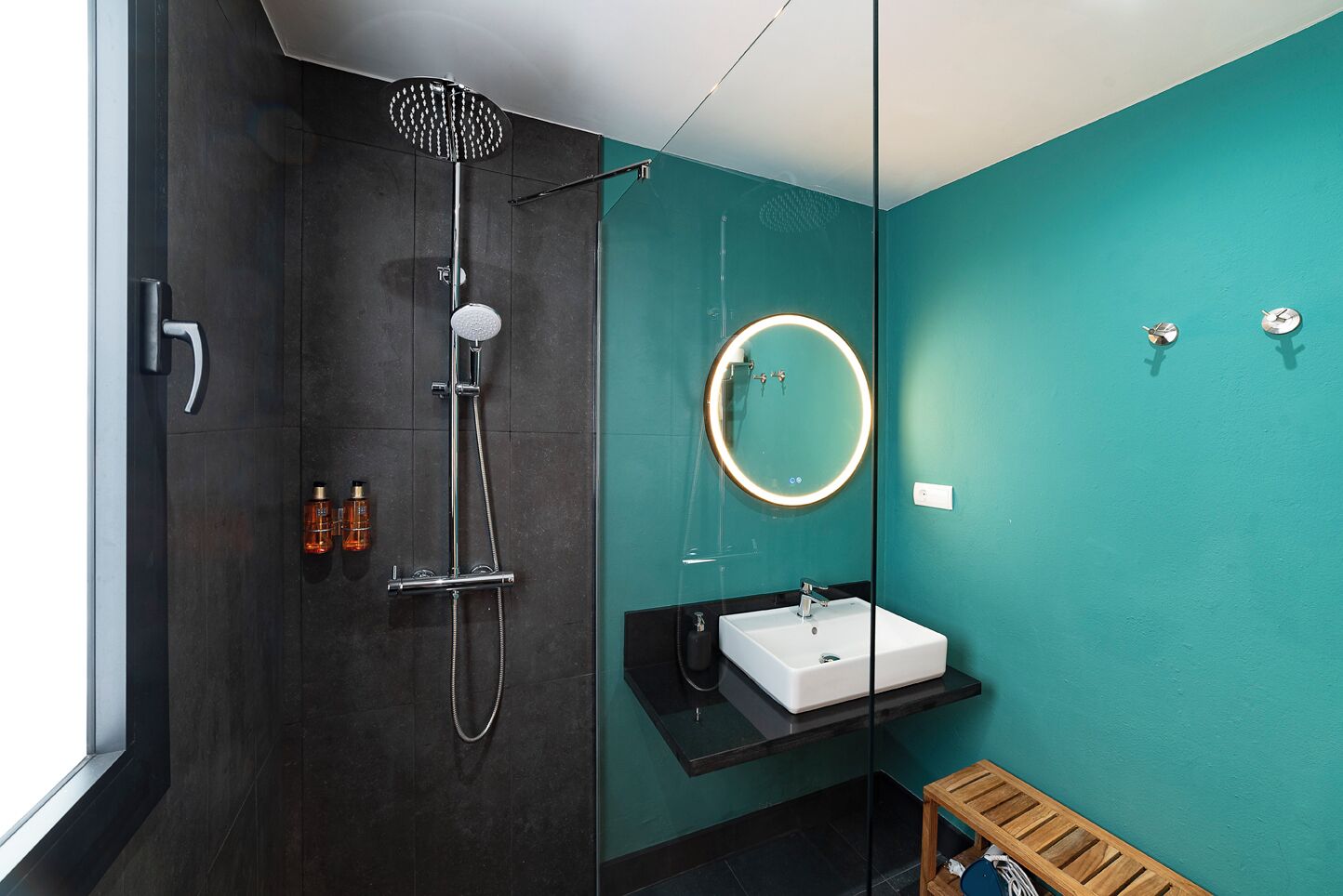 A modern bathroom with a dark shower, a white sink, a round mirror, and a teal wall.