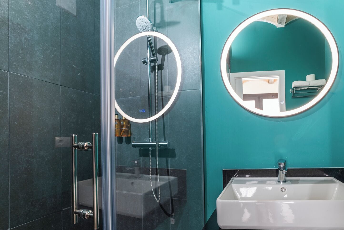 A modern bathroom with a glass-enclosed shower, a round mirror with a light, and a white sink.