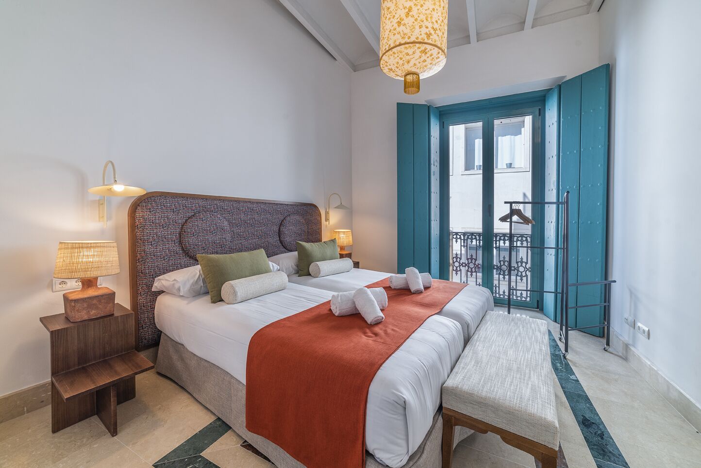 Quarto de hotel com uma cama grande, duas mesas de cabeceira, um banco, uma varanda com guarda-corpo e vista para o exterior.