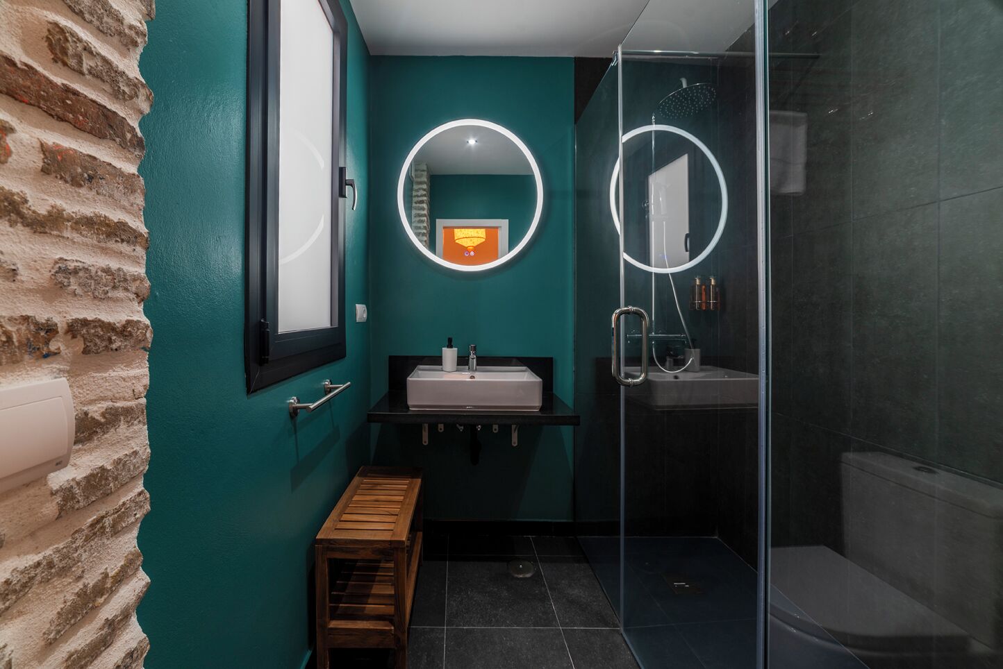 Een badkamer met een blauwgroene wand, een houten bankje, een wastafel met spiegel en een douchecabine van glas.