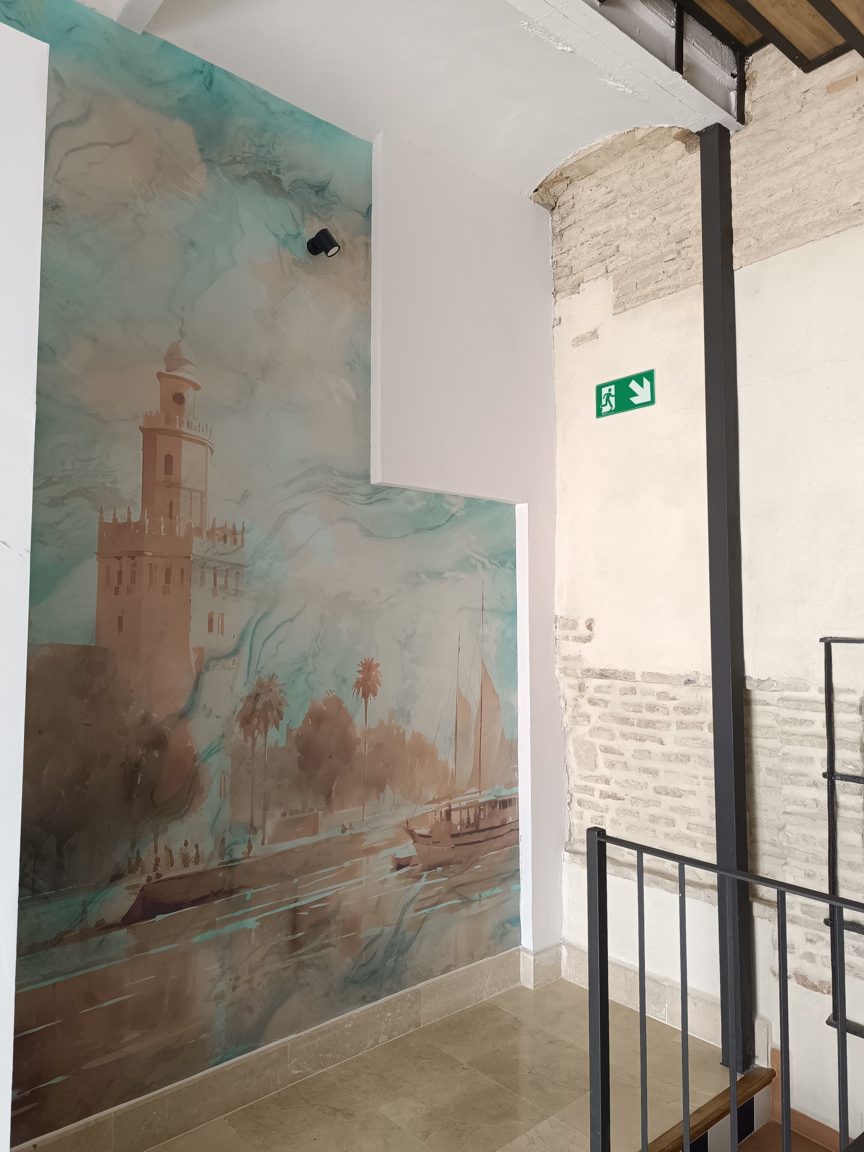 A corridor with a mural of a coastal scene, a brick wall, and a green emergency exit sign.