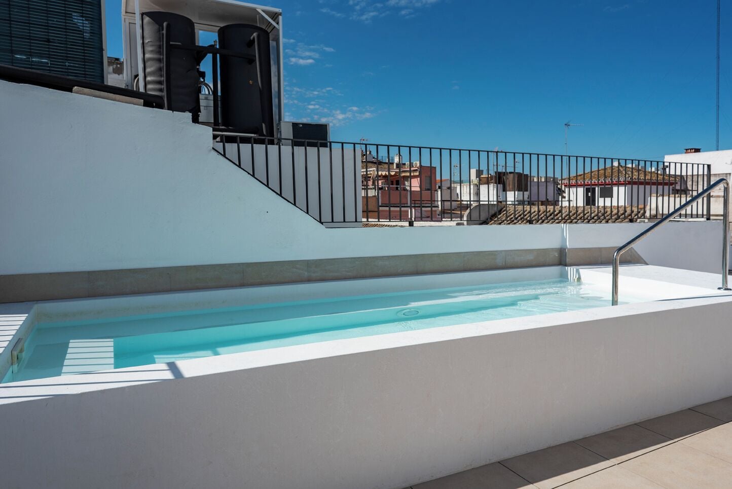 A rooftop swimming pool with a metal railing, a staircase, and buildings in the background.