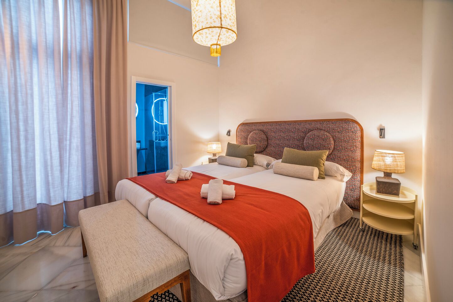 A hotel room with a large bed, two bedside tables, a bench, a patterned rug, and a chandelier.