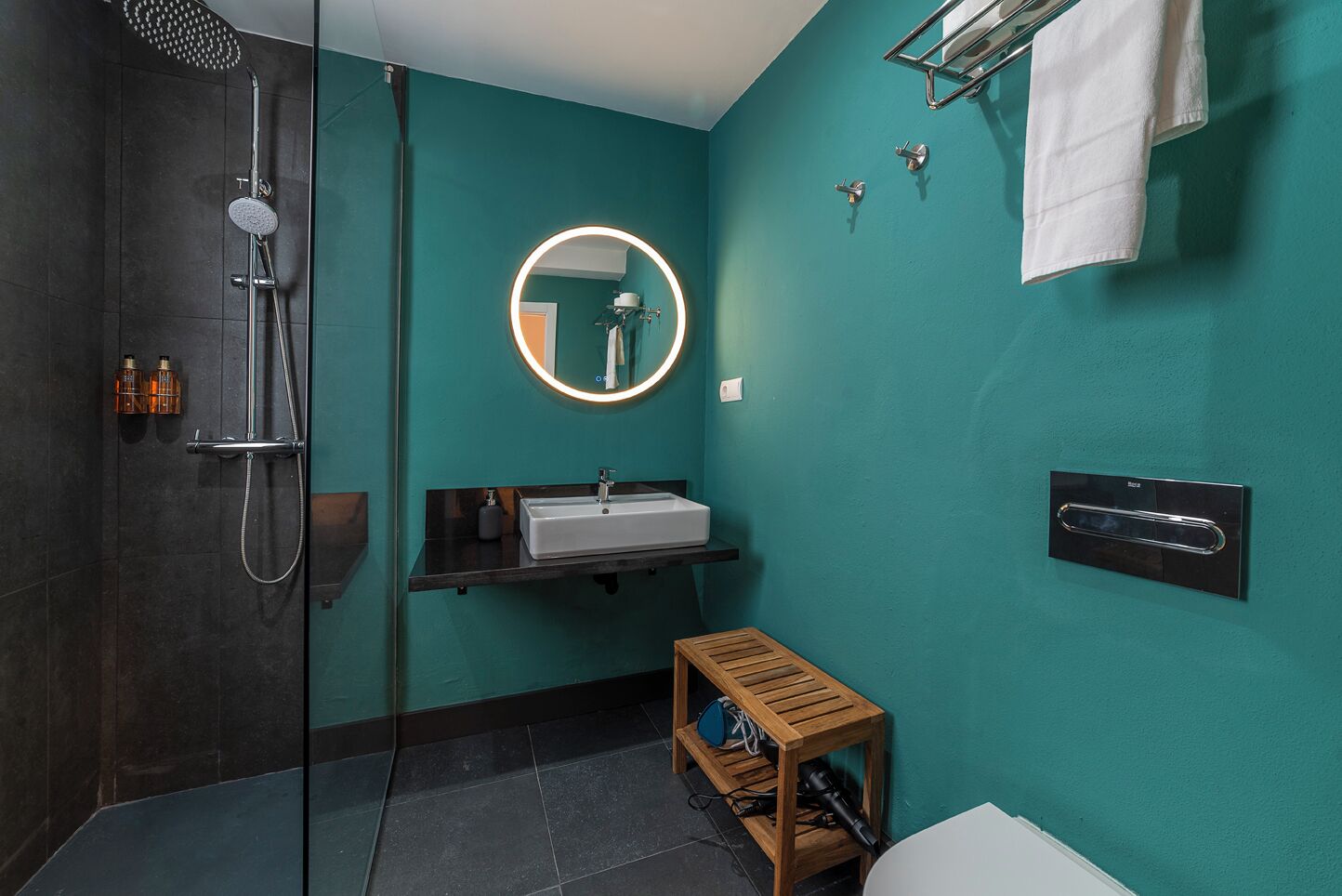 Uma casa de banho moderna com chuveiro em cabine de vidro, espelho redondo com luz, lavatório branco e um banquinho de madeira.