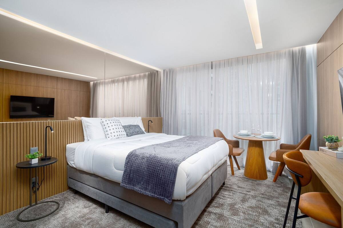 A modern hotel room with a bed, bedside table, TV, and a dining area with chairs and a table.