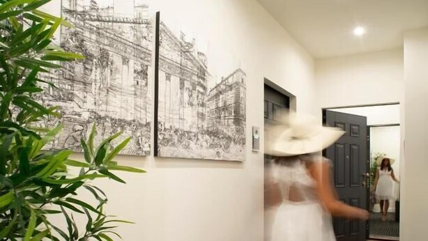 Un pasillo con obras de arte blanco y negro en las paredes, una planta en maceta y una persona que camina por el corredor con un vestido y sombrero blancos.