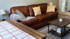 A living room with a brown leather sofa, a wooden coffee table, a plaid tablecloth, a wall clock, and a door with frosted glass panels. - Cozy basement apartment suite, beautiful Paradise! (Paradise)