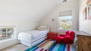 A bedroom with a bed, a colorful striped rug, a red chair, and a wooden cabinet. - Heated Pool Grill Breakwater by Rovetravel (Sag Harbor)