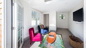 A modern living room with a large sliding glass door, a flat-screen TV mounted on the wall, and a colorful rug. - Heated Pool Grill Breakwater by Rovetravel (Sag Harbor)