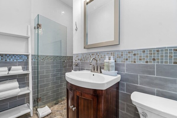 A bathroom with a glass shower enclosure, a white sink with a wooden vanity, and a large mirror above the sink. - Heated Pool Beach Miramar Villa by Rovetravel (West Palm Beach)