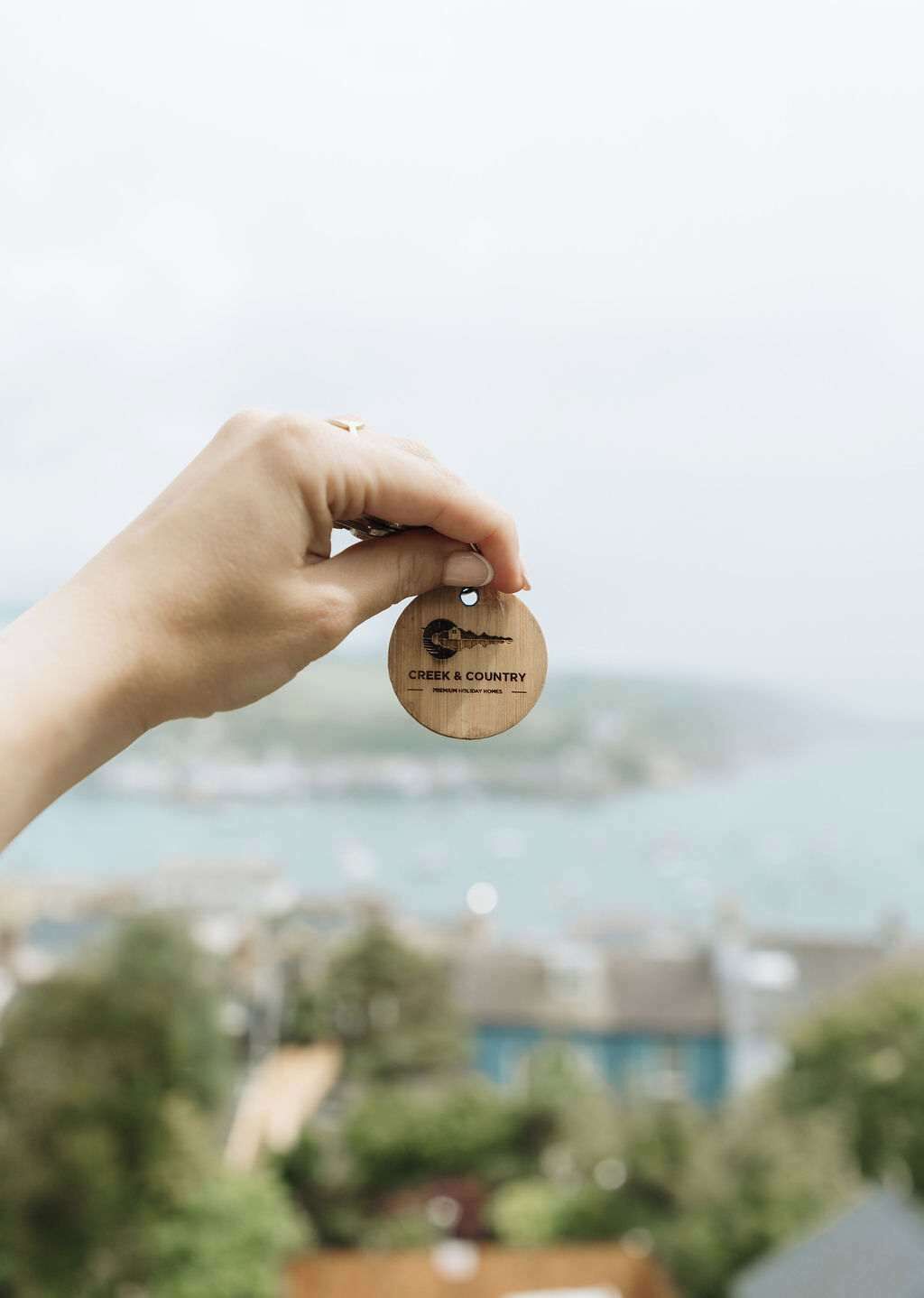 A hand holding a wooden keychain with 'CREEK & COUNTRY' engraved on it, set against a blurred background of a coastal cityscape.