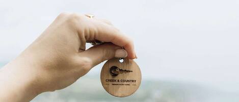 A hand holding a wooden keychain with 'CREEK & COUNTRY' engraved on it, set against a blurred background of a coastal cityscape.