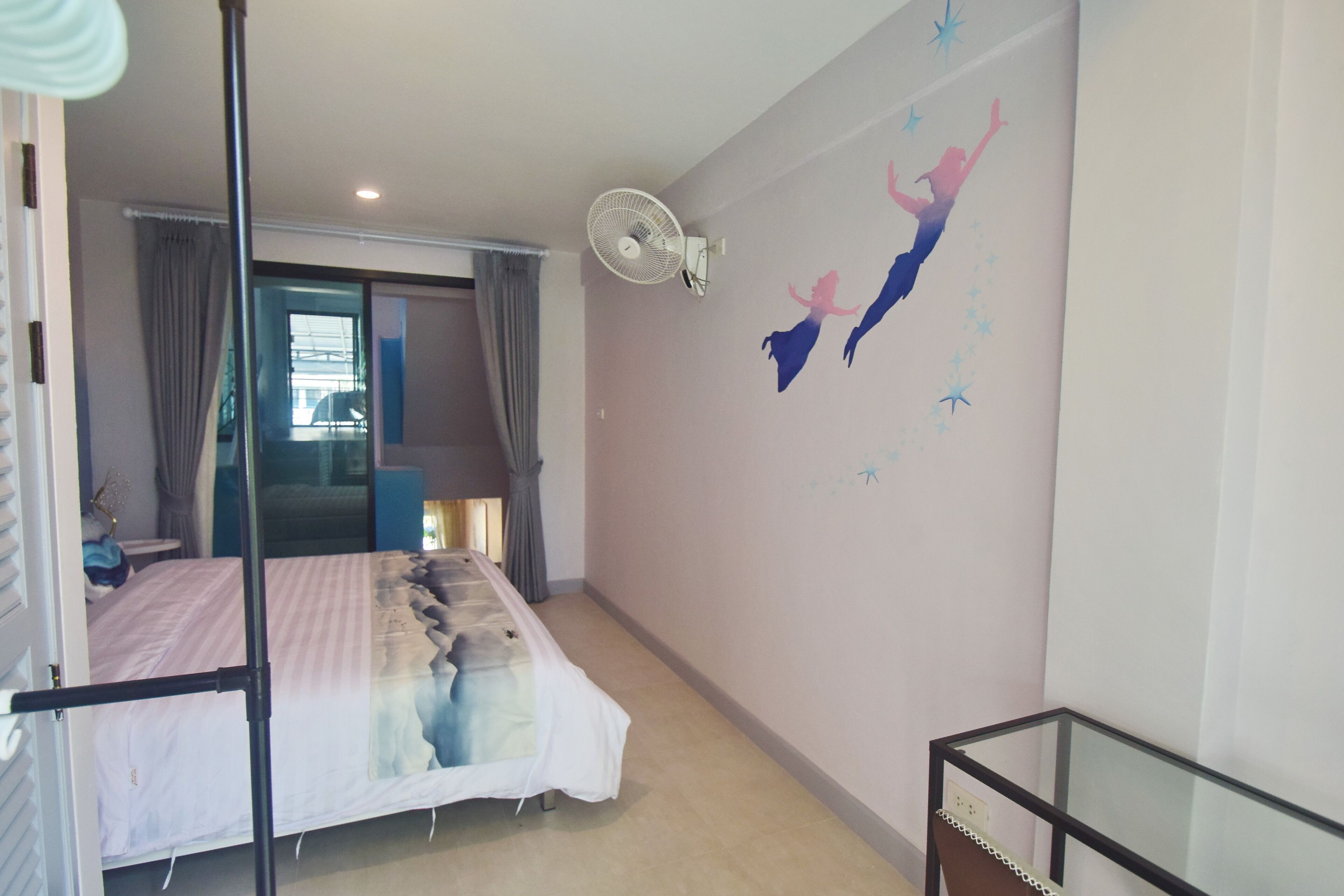 Ein Schlafzimmer mit einem Bett, einem Standventilator, einem Glastisch und Wandbild.
