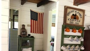 Un hall d’entrée accueillant avec une porte en bois, une étagère verte ornée de tasses blanches, un drapeau américain suspendu et une enseigne publicitaire ancienne pour le café.