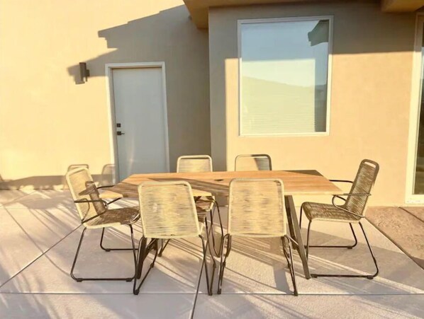 Un tavolo rettangolare in legno con sei sedie intrecciate, posizionato su un patio piastrellato, di fronte a un muro beige con una porta e una finestra.
