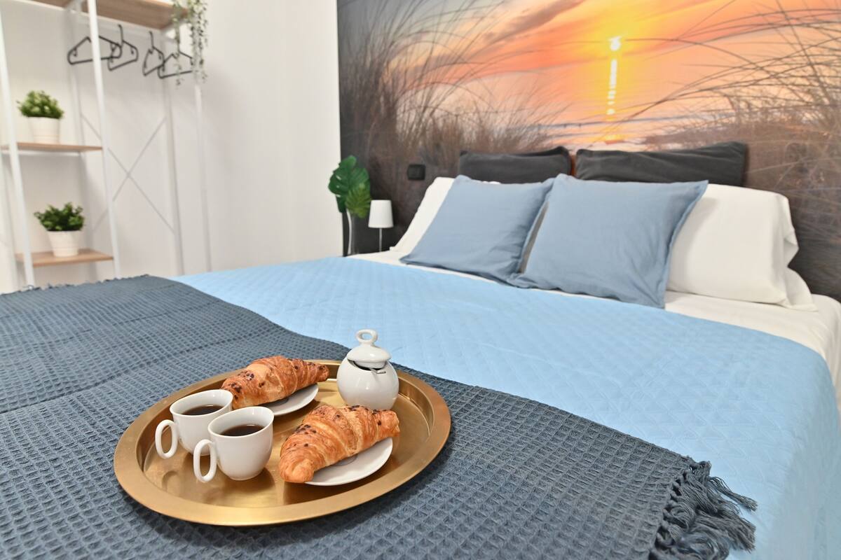 A neatly made bed with a blue blanket, a tray with croissants and coffee cups, and a painting of a sunset on the wall.