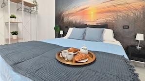 Una cama bien hecha con una colcha azul, una bandeja con croissants y café, y un cuadro en la pared de una puesta de sol.