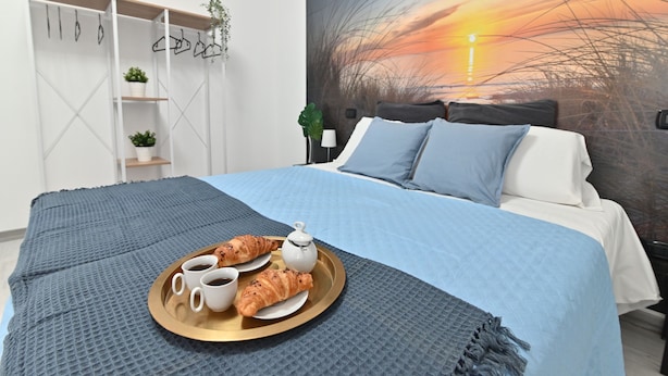 Una cama bien hecha con una manta azul, una bandeja con croissants y café, y un cuadro de atardecer en la pared.