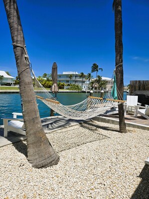 A hammock between palm trees by a pool with a clear view of a building and a boat.
