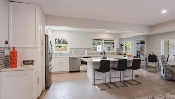 Une cuisine moderne avec des meubles blancs, un îlot central doté de tabourets de bar et une vue sur l’extérieur.