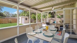 Une terrasse couverte avec une table préparée pour le petit-déjeuner, des chaises blanches et un ventilateur de plafond.