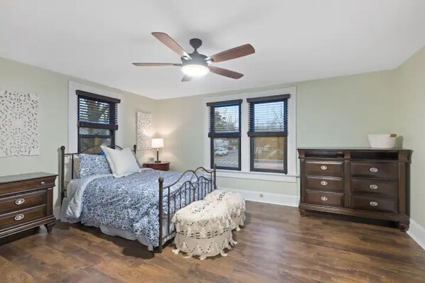 Un dormitorio con cama, mesitas de noche, cómoda, ventilador de techo y ventanas amplias.