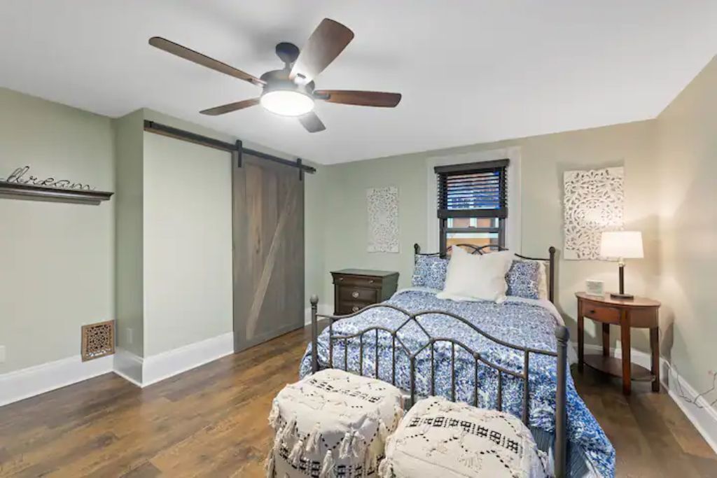 Ein Schlafzimmer mit Bett, Deckenventilator, Fenster mit Jalousien, Nachttisch und Beistelltisch.