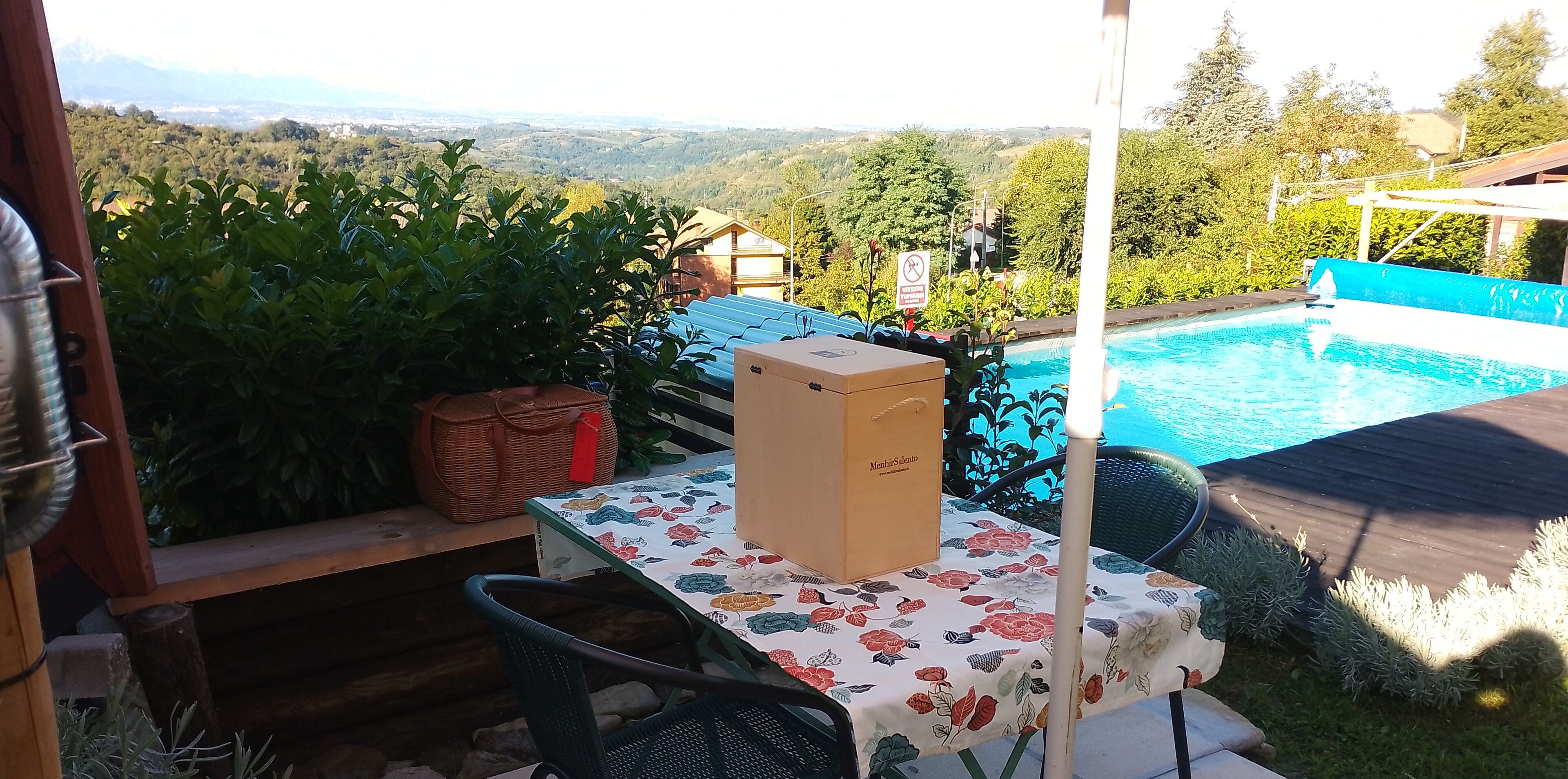 Uma área de pátio com uma mesa, cadeiras, uma cesta de vime e uma caixa com a etiqueta 'Mehlsilento'. Uma piscina é visível ao fundo.