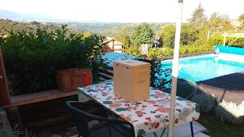 Uma área de pátio com uma mesa, cadeiras, uma cesta de vime e uma caixa com a etiqueta 'Mehlsilento'. Uma piscina é visível ao fundo.