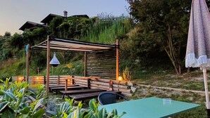 Ein rustikaler Außenbereich mit Holzbänken und -tischen, einer Pergola mit hängenden Lichtern und Blick auf einen Hügel mit einem Gebäude im Hintergrund.