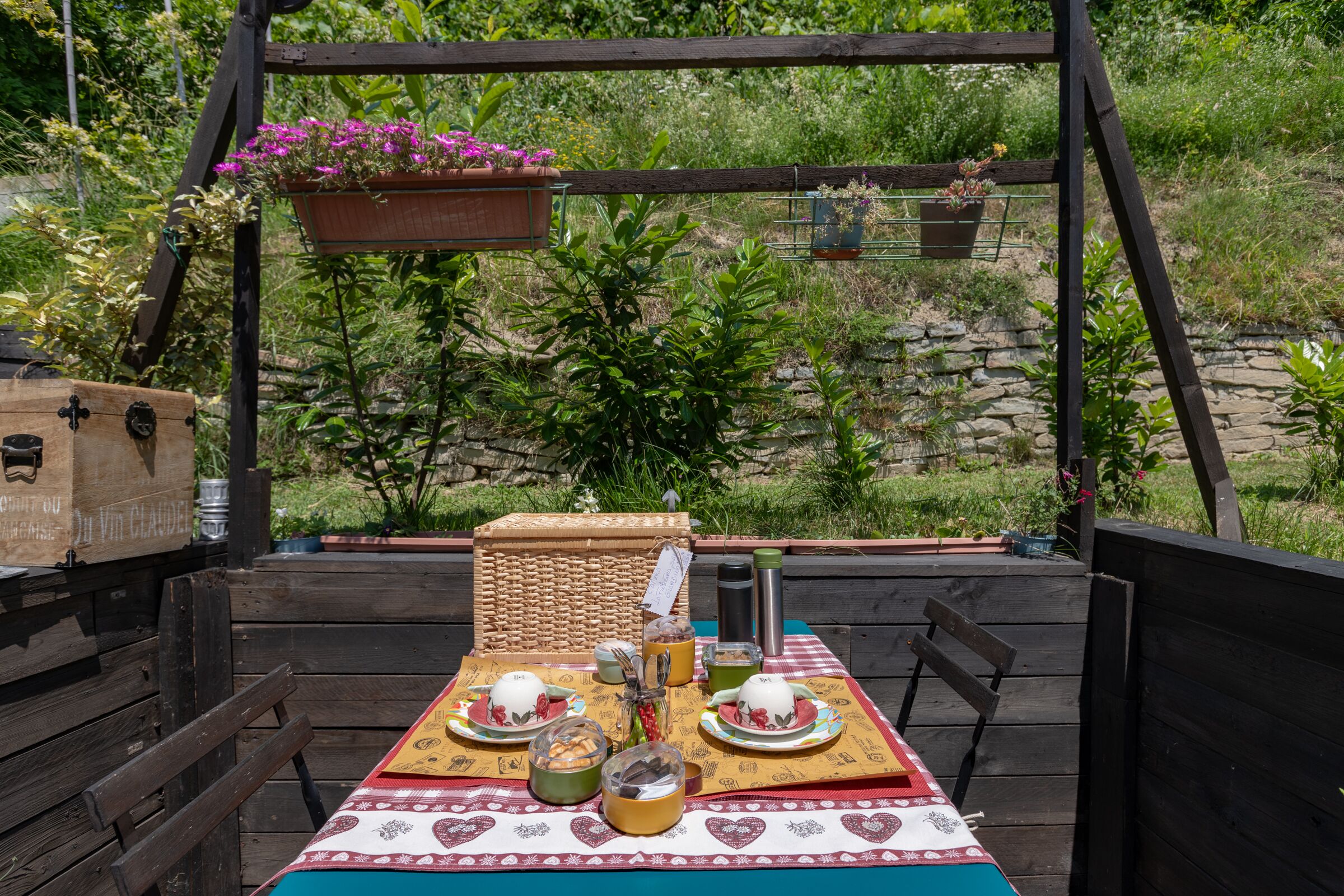An outdoor dining area with a wooden table set for two, surrounded by greenery and a stone wall.