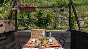 An outdoor dining area with a wooden table set for two, surrounded by greenery and a stone wall.