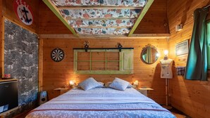 Une pièce chaleureuse aux murs de bois, avec un lit, un jeu de fléchettes, un miroir et un mur orné d’un motif floral.
