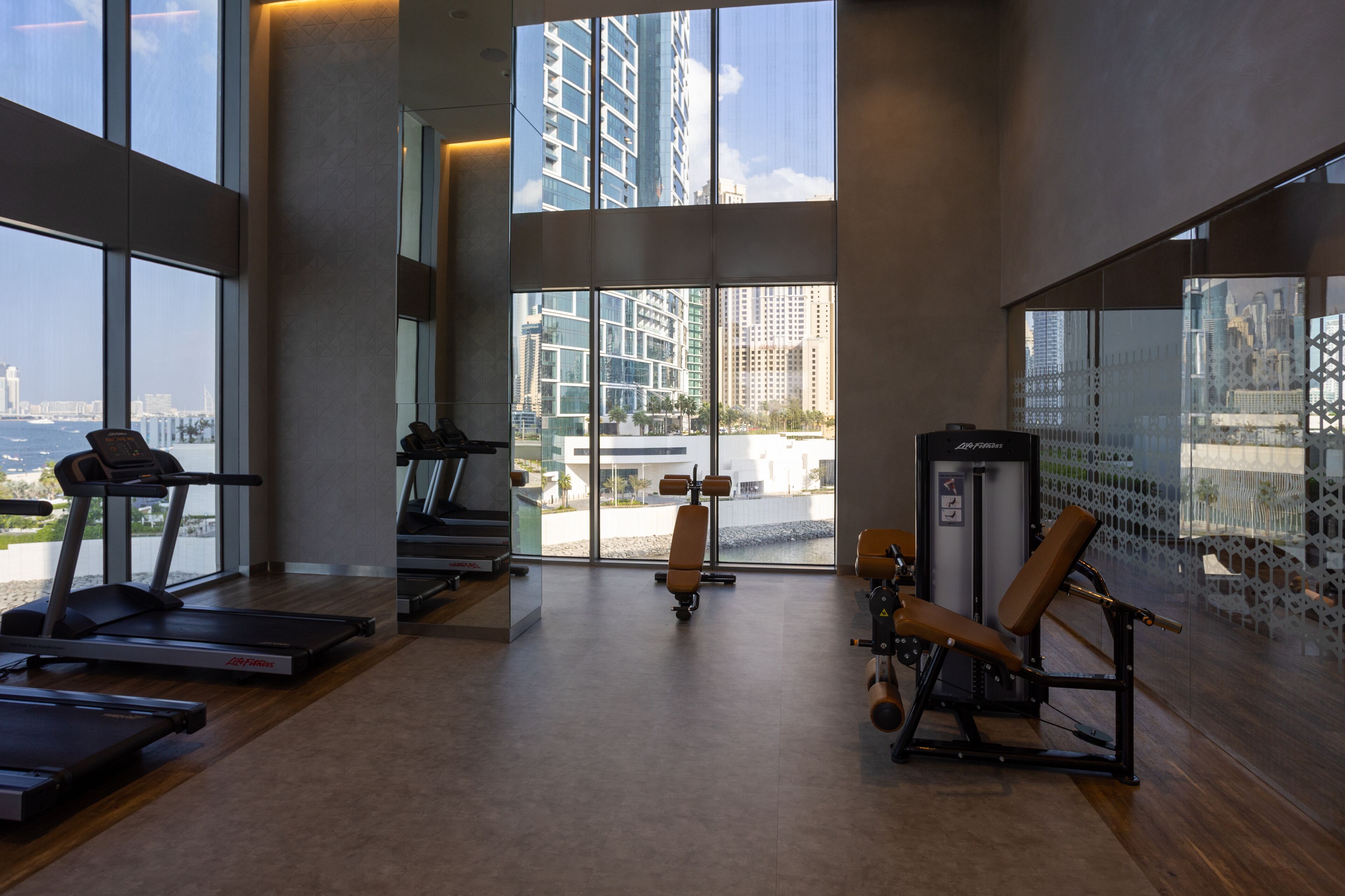 A gym with treadmills, a stationary bike, and weight machines, with large windows offering views of a cityscape.
