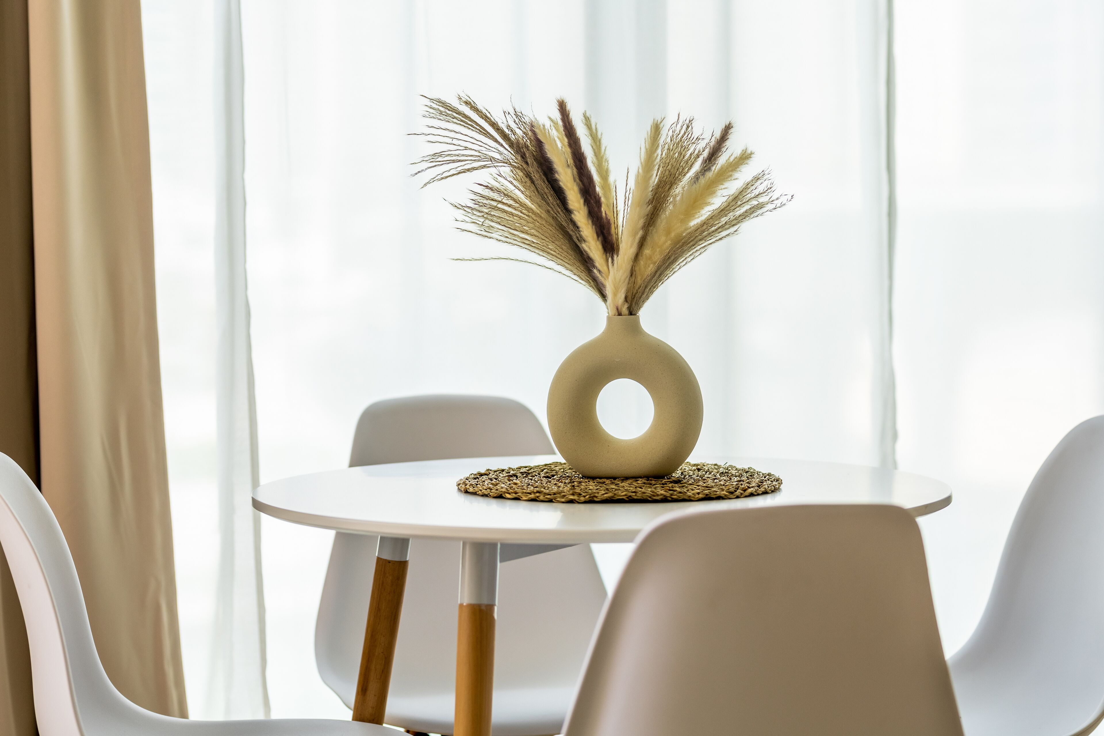 Une table ronde blanche, sur laquelle est posé un vase contenant des plantes séchées, est entourée de chaises blanches aux pieds en bois, dans une pièce où sont accrochées des rideaux légers et transparents.