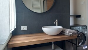 A modern bathroom with a wooden vanity, a white sink, a round mirror, and a built-in washing machine. - Casa Maria, Nuovo Appartamento con Vista Lago (Albisano di Torri del Benaco)