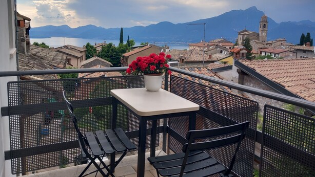 A balcony with a table and chairs, overlooking a mountainous landscape with a lake and buildings. - Casa Maria, Nuovo Appartamento con Vista Lago (Albisano di Torri del Benaco)