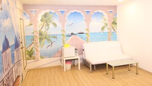 Una habitación con un mural de una playa, un sofá blanco, una mesita con una lámpara y un cubo de basura.