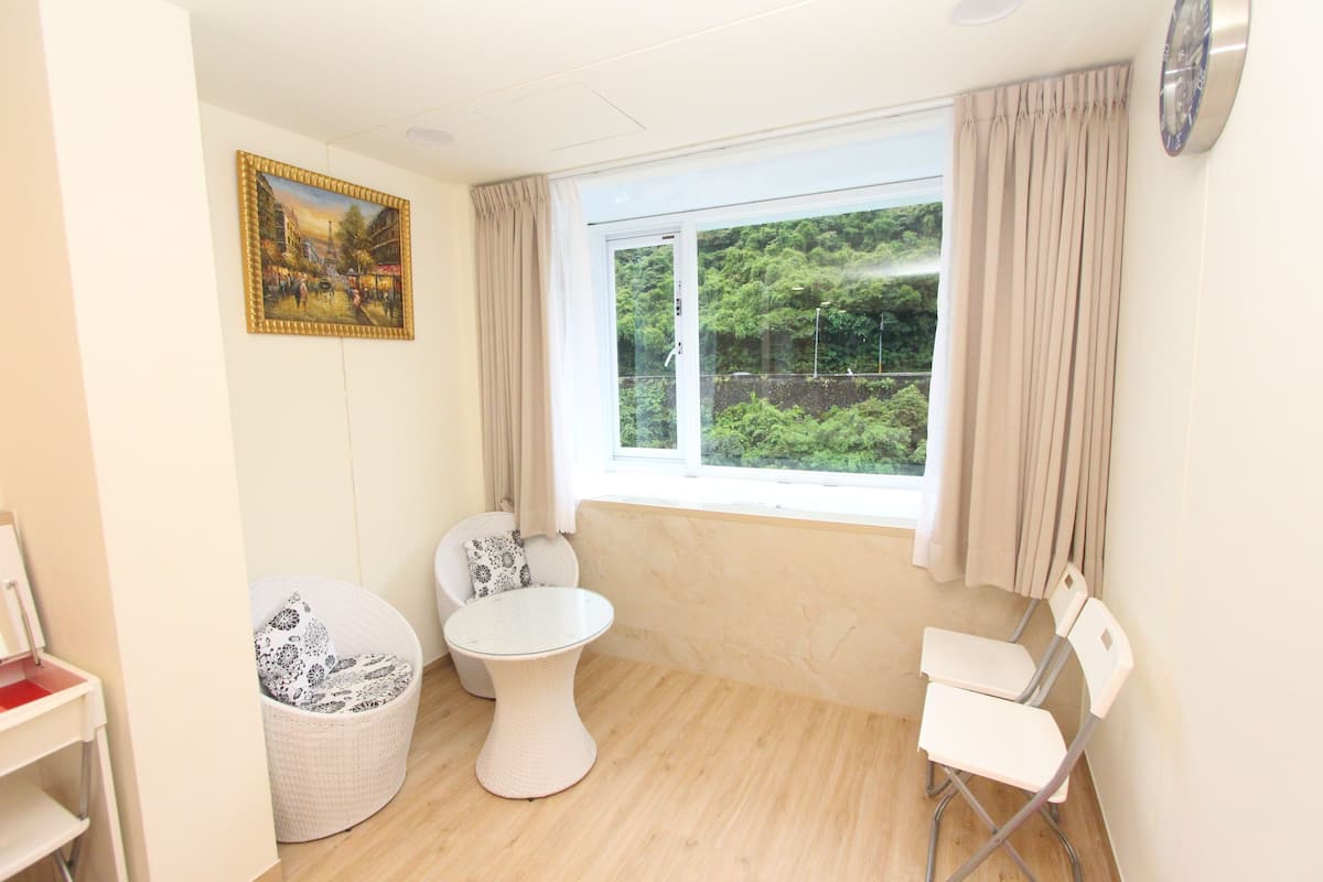 Una habitación con un ventanal grande, un cuadro en la pared, una silla blanca, una mesita pequeña y una silla de mimbre.